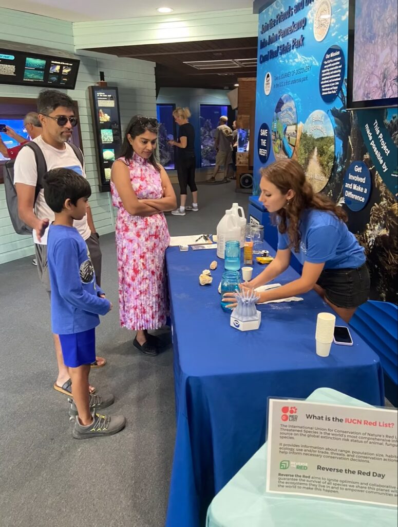 A Coral Restoration Foundation educator demonstrates a hands-on coral science activity at an indoor exhibit table, engaging a family with a child while explaining reef conservation concepts using coral samples, jars of seawater, and educational materials in a museum-style learning space