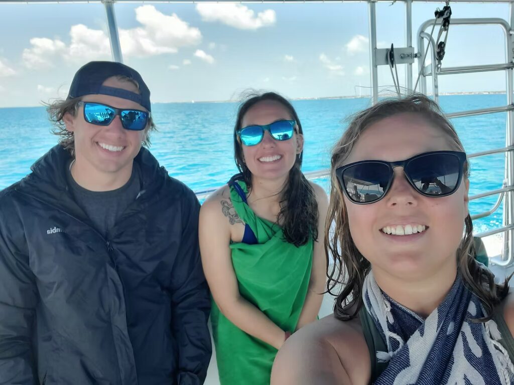 Three smiling people wearing sunglasses take a selfie on a boat, with bright blue ocean and a sunny sky behind them.