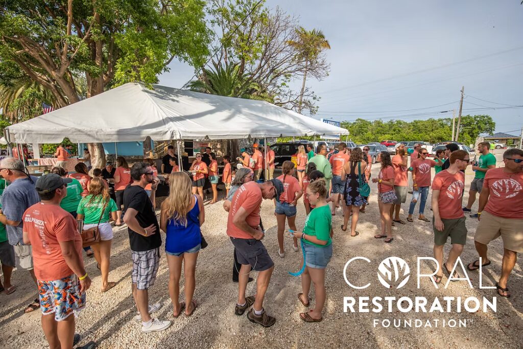 A large crowd of Coralpalooza participants in colorful event T-shirts gather under and around a white event tent for check-in or festivities, with the Coral Restoration Foundation logo displayed in the corner.