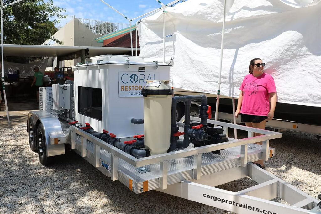 A white mobile coral transport and life-support trailer sits on a gravel lot. It has the Coral Restoration Foundation logo on the side and is equipped with pumps, pipes, and filtration units. A woman in a bright pink shirt and sunglasses stands nearby, looking at the trailer.