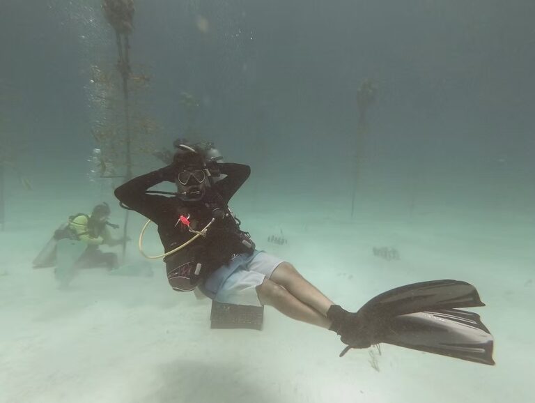 A scuba diver relaxes underwater near coral nursery trees, floating with legs crossed and hands behind their head while another diver works in the background.