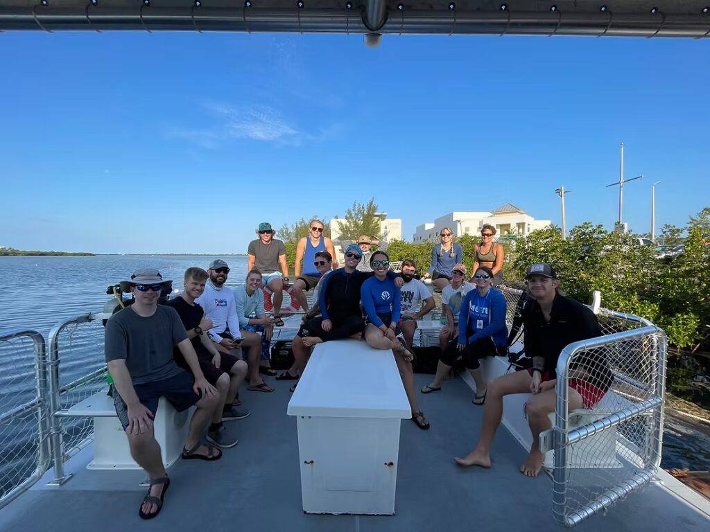 A large group of people sit together on the deck of a dive vessel before heading out to a restoration site, posing for a group photo with calm water and mangroves behind them.