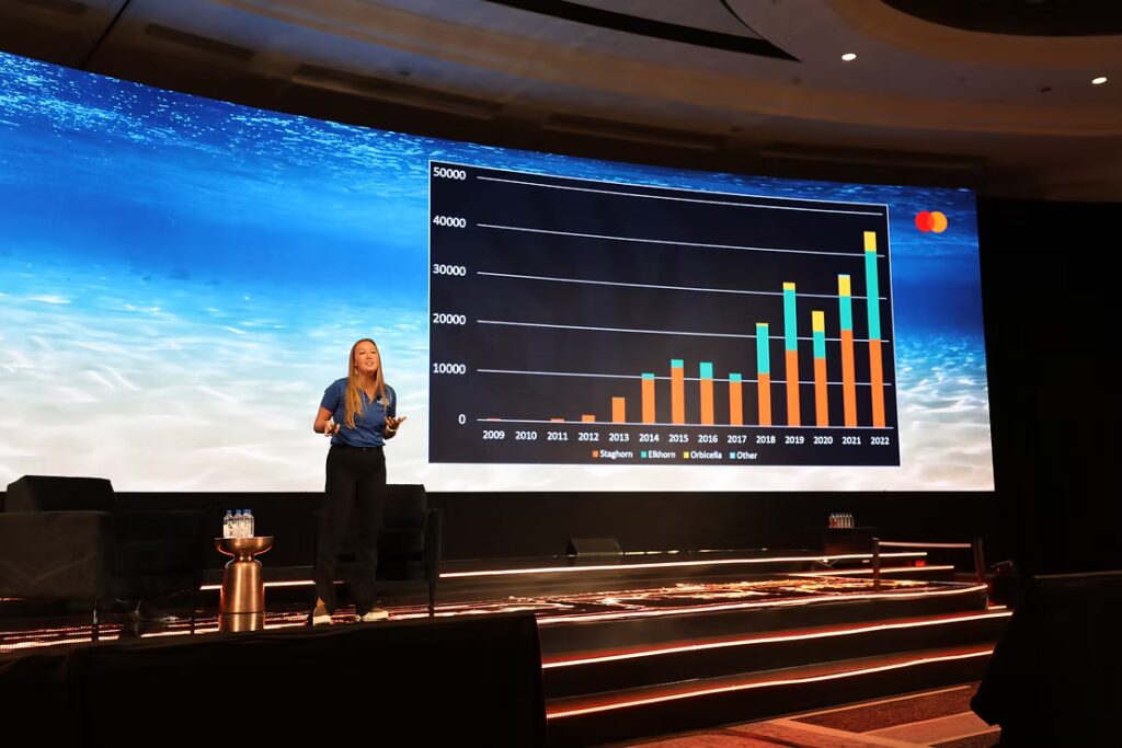A presenter stands on a stage in front of a large screen showing a bar graph of coral outplant numbers by species from 2009 to 2022, with an ocean background projected behind her.