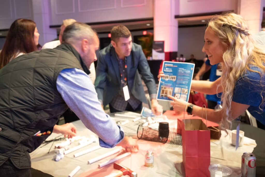 Participants gather around a table assembling coral nursery materials. A woman on the right explains from an instruction sheet while others work with PVC pipes and mesh.
