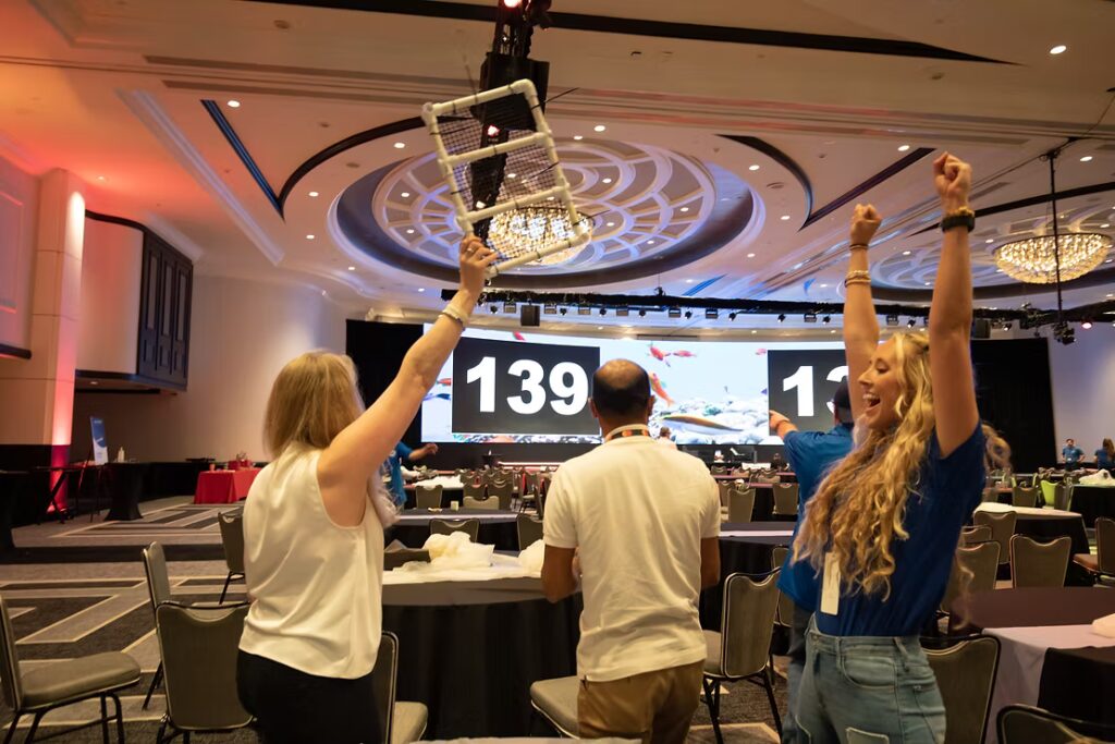 Three people cheer inside a large conference room, one holding up a PVC Coral Tree frame. A giant screen behind them displays numbers in a competitive team-building setting.
