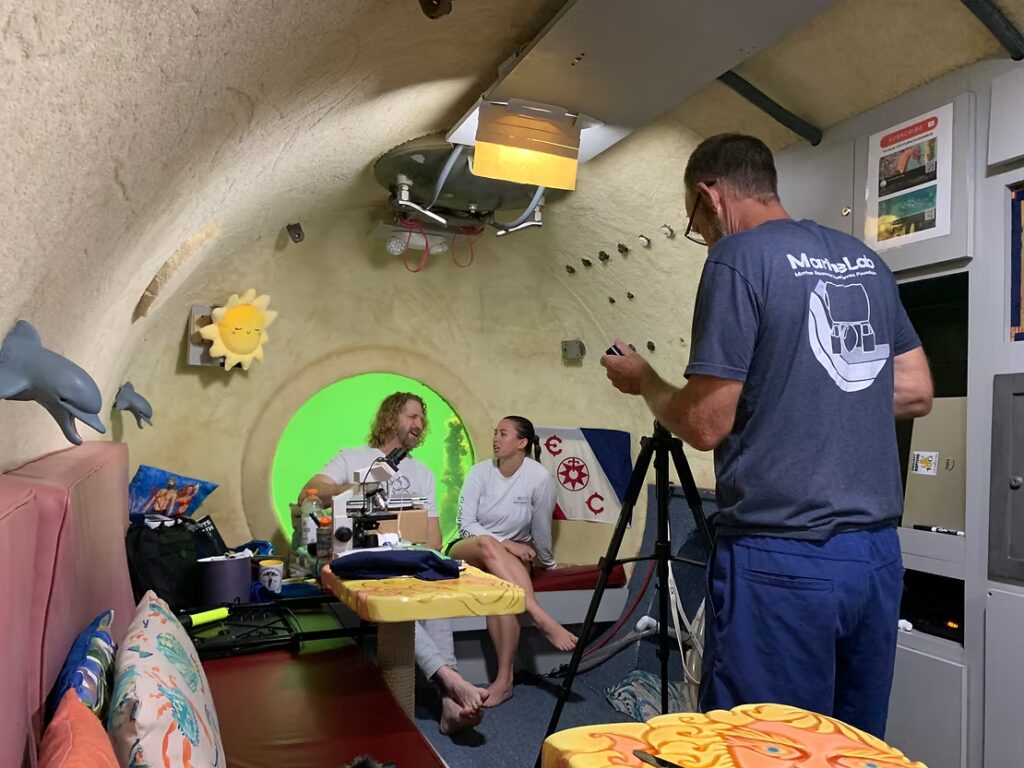 Inside an underwater research habitat, a CRF staff member is interviewed at a small table while another person films with a tripod, with colorful marine-themed decor on the curved walls.