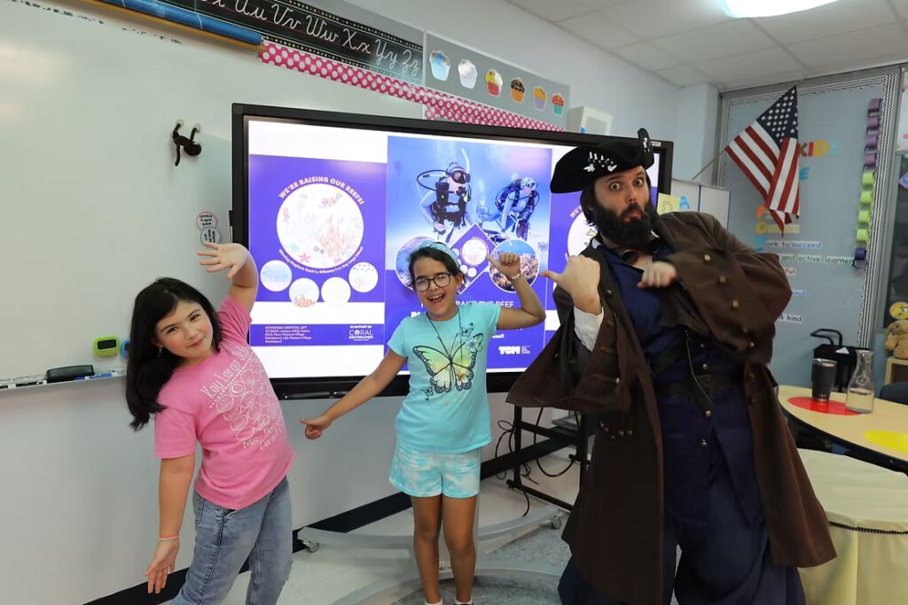 A Coral Restoration Foundation educator dressed as a pirate poses playfully with two elementary school students in front of a large classroom screen showing coral restoration graphics. The children smile and strike fun poses, highlighting an engaging classroom outreach program.