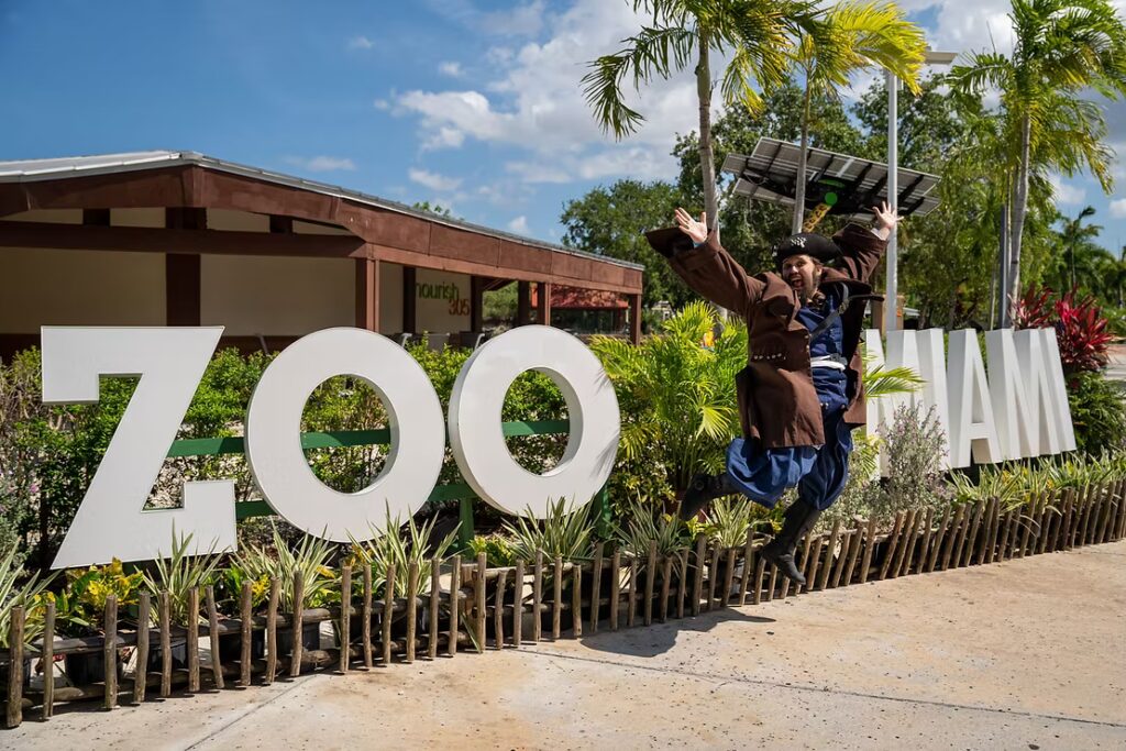 A costumed pirate jumps enthusiastically into the air with arms spread wide in front of the large white “ZOO MIAMI” sign. Tropical plants and bright blue sky fill the background.