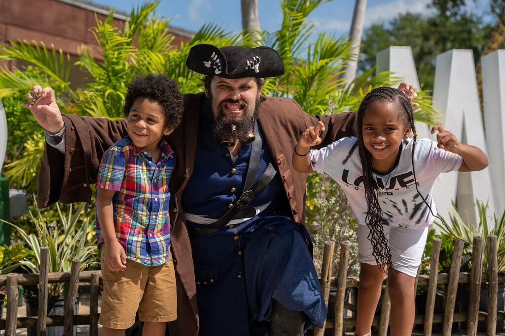 A costumed pirate character crouches playfully with two smiling young children on either side, all making silly “claw” poses. They stand outdoors in front of lush green plants on a sunny day.