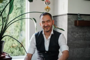 Photo of Indigo Dandelion Lee Mané wearing a white button up and vest while sitting in front of window