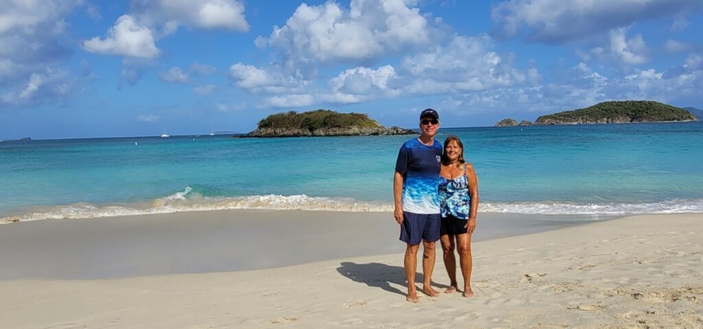 A man and a woman standing together on a beach with Caribbean islands and cerulean water behind them.