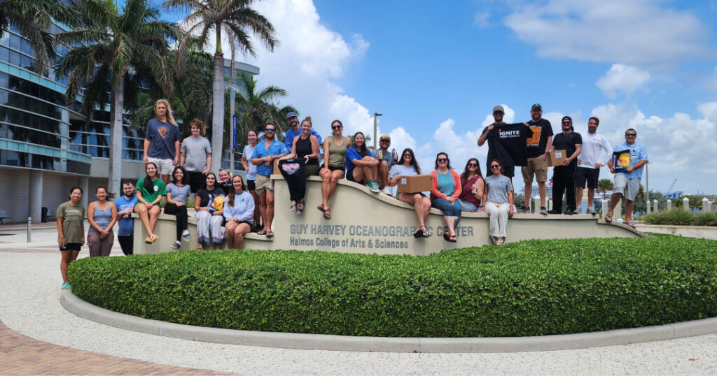 Large group of coral restoration practitioners from various organizations smiling and gathered around the Guy Harvey Oceanographic Center sign at Nova Southeastern University, under palm trees and a blue sky.