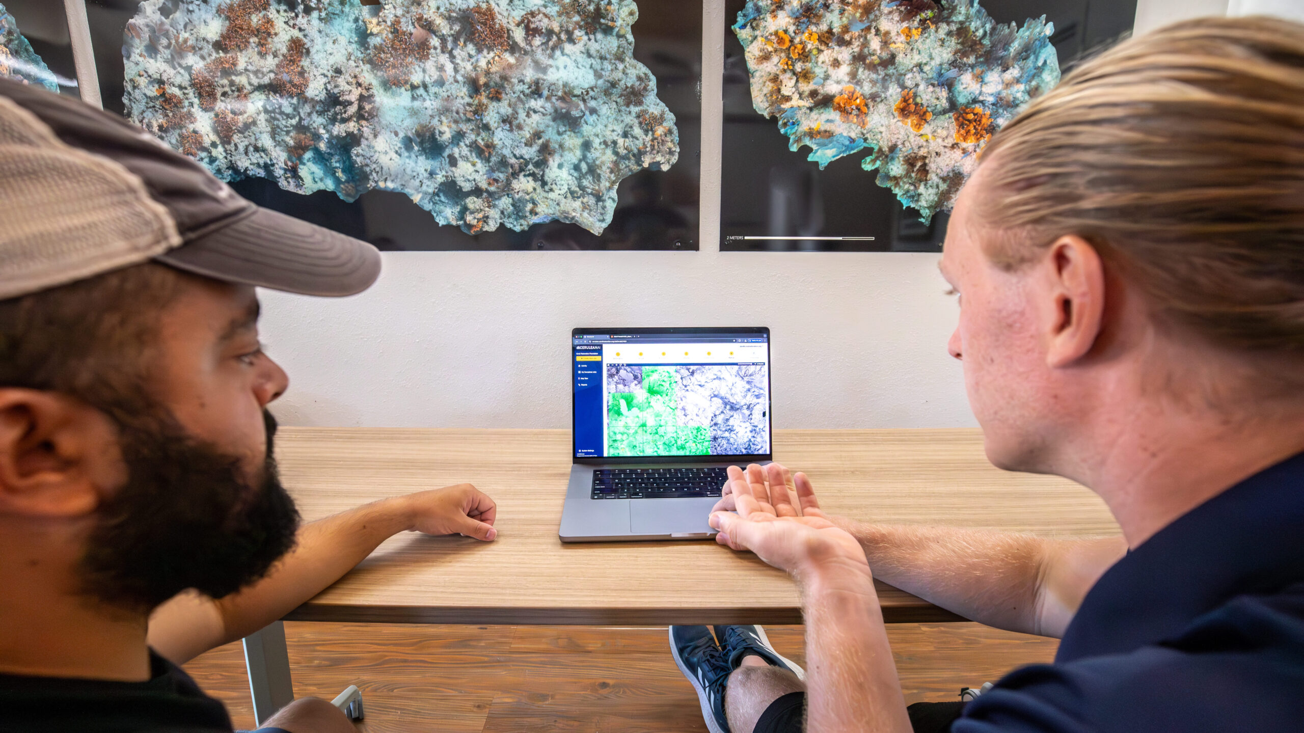 Two team members reviewing coral photomosaic imagery on a laptop at a desk, with large reef photomosaics displayed on the wall in the background.