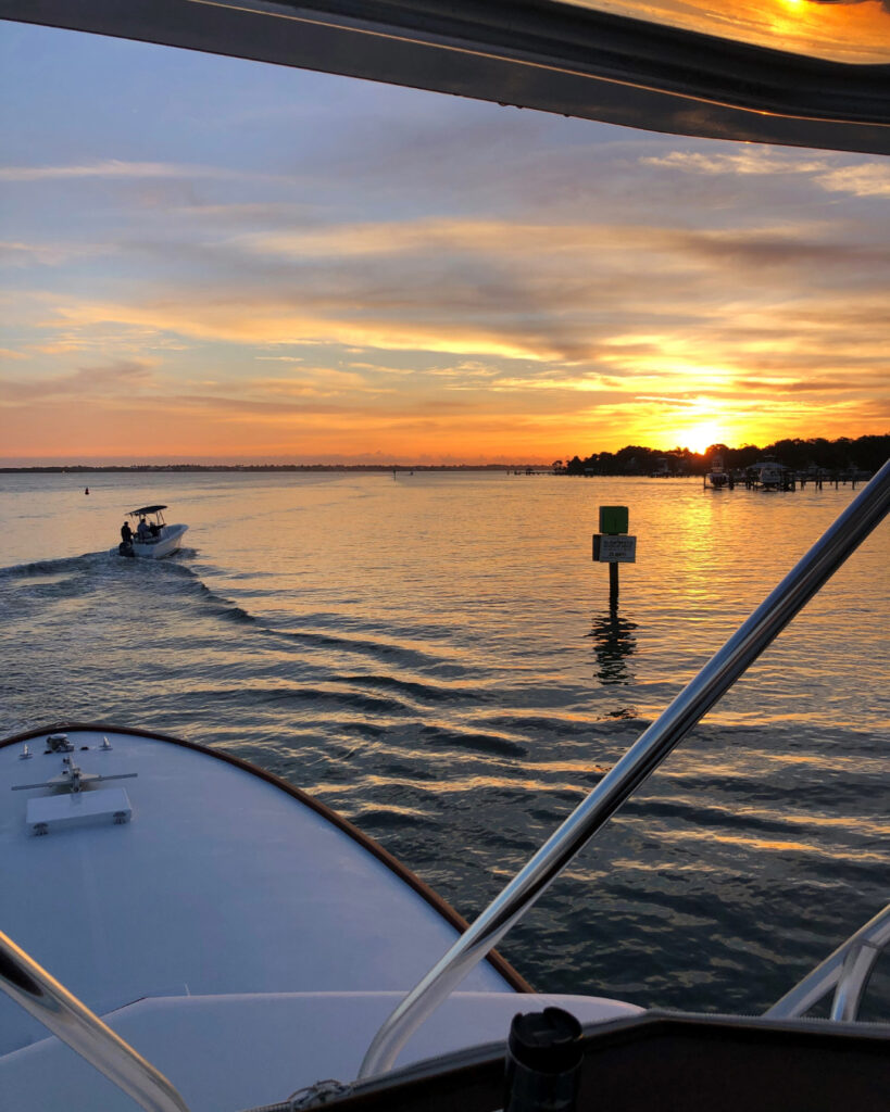 A boat cruises across calm waters at sunrise, viewed from another boat’s bow, with the sun rising over the shoreline.
