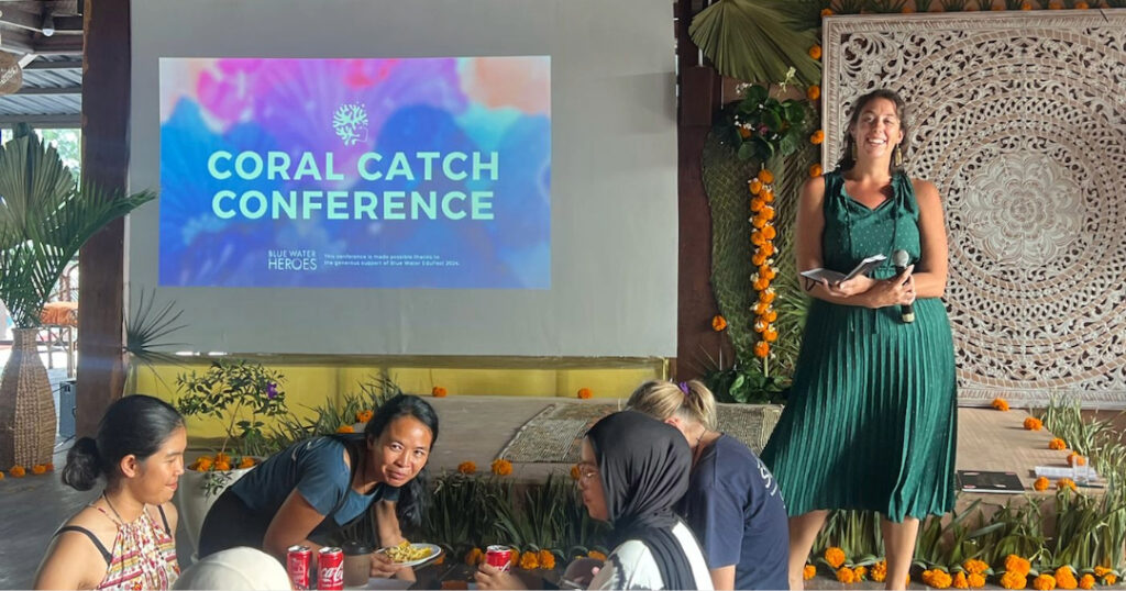 CRF Director of Restoration Strategy Jessica Levy stands on stage at the Coral Catch Conference in Gili Air, Indonesia, smiling as she speaks to attendees seated below a screen displaying the event title