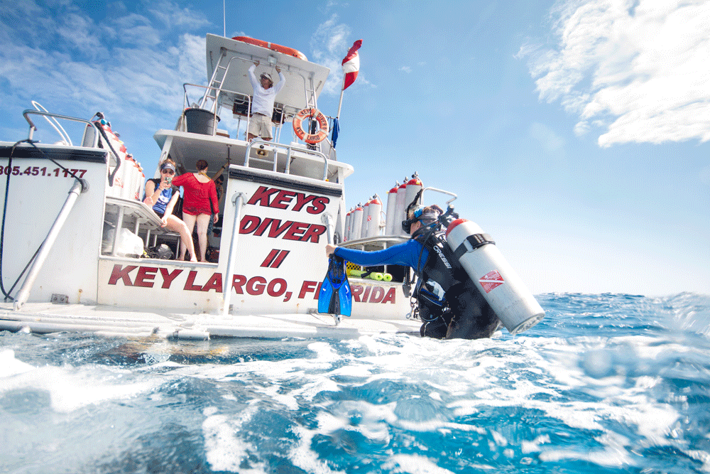 A scuba diver carrying a tank enters the ocean from the Keys Diver II boat in Key Largo, Florida, while other divers prepare onboard under sunny skies.