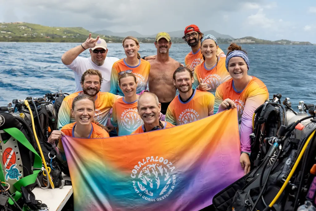 A group of smiling divers in matching sun shirts poses on a boat, holding a vibrant CoralPalooza banner after a day of planting coral.
