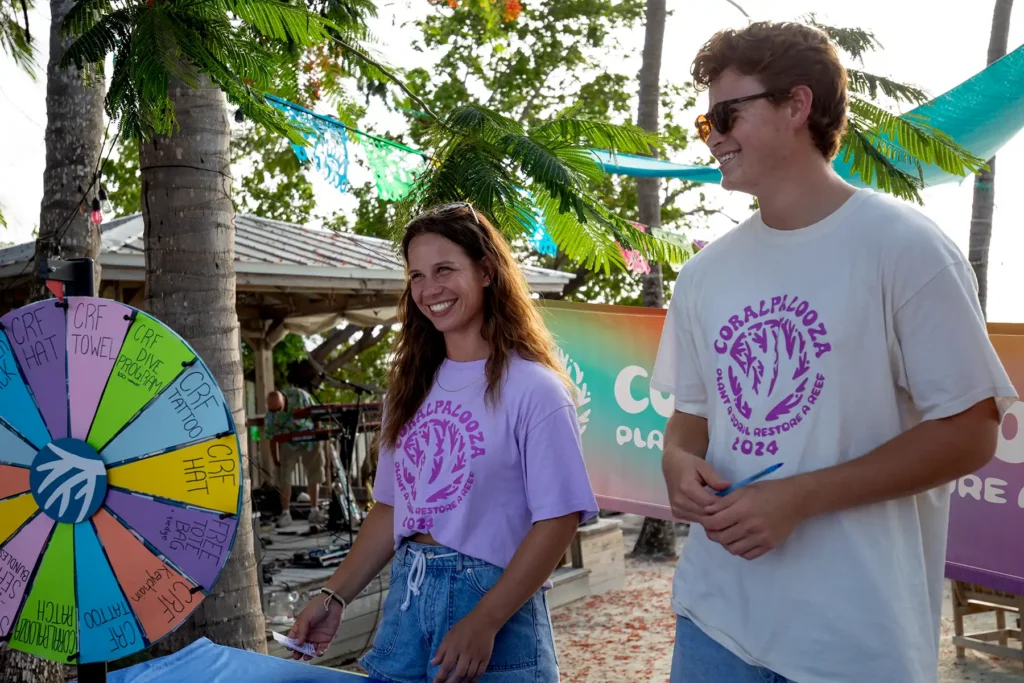 Two volunteers in CoralPalooza t-shirts laugh next to a colorful prize wheel at a tropical outdoor reef restoration awareness event.