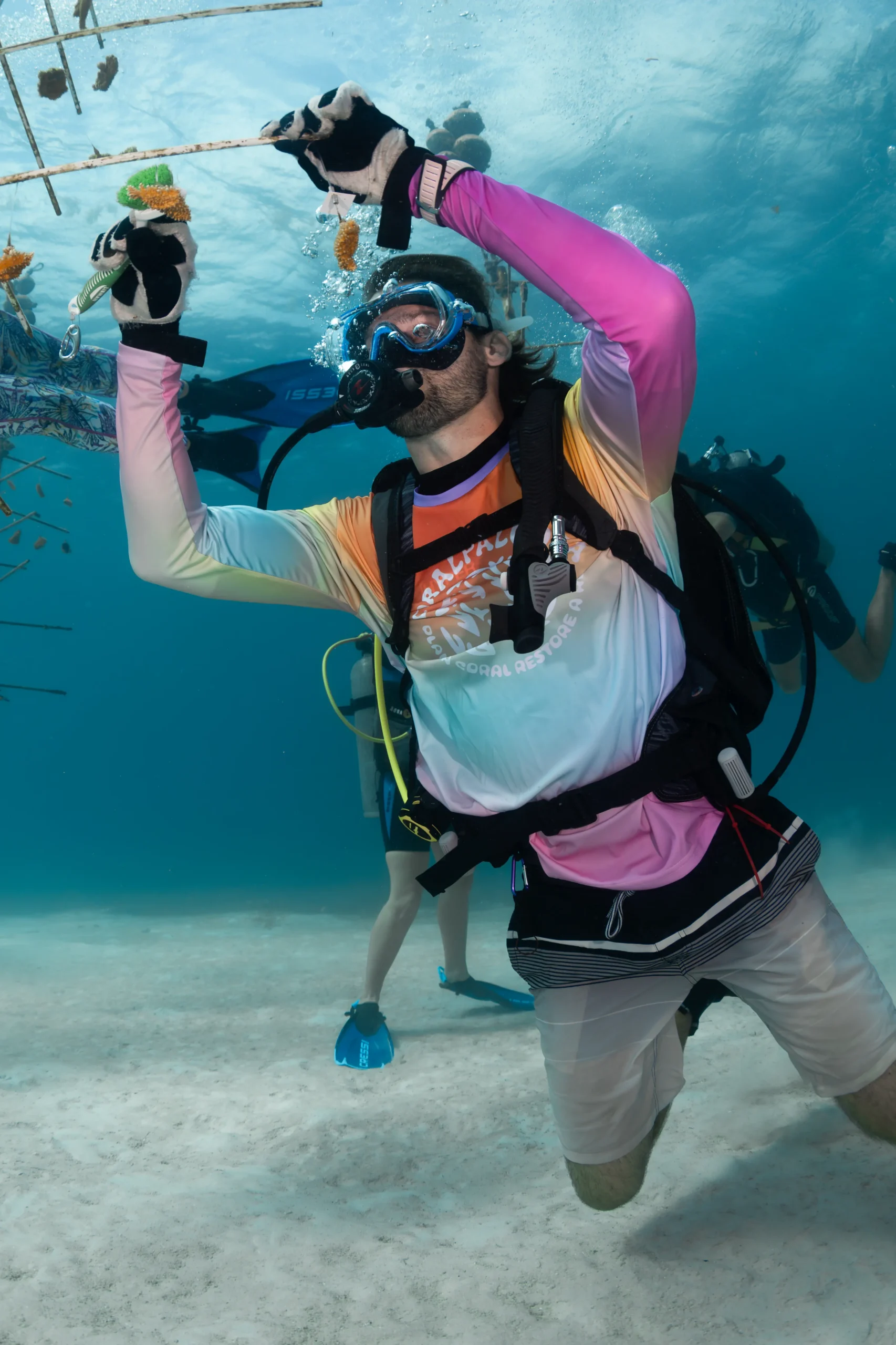 A scuba diver in a blue rash guard examines brain coral on the ocean floor while bubbles rise toward the surface in a clear tropical reef environment.